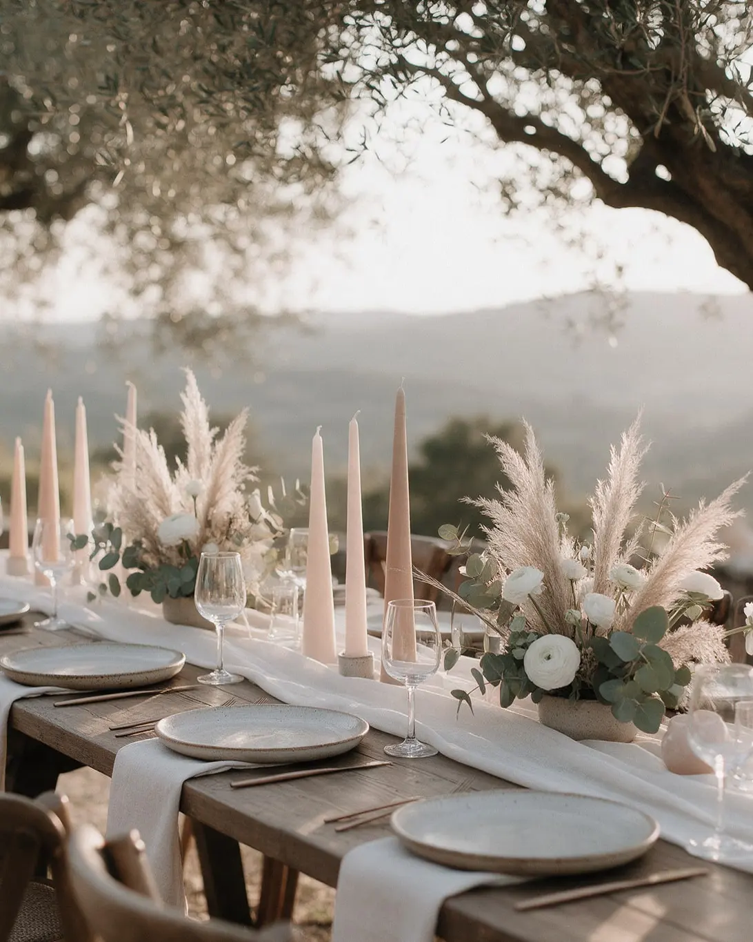 Longue table de mariage sous des oliviers en Provence, chemin de table en lin ivoire, bougies rose poudré et pampas