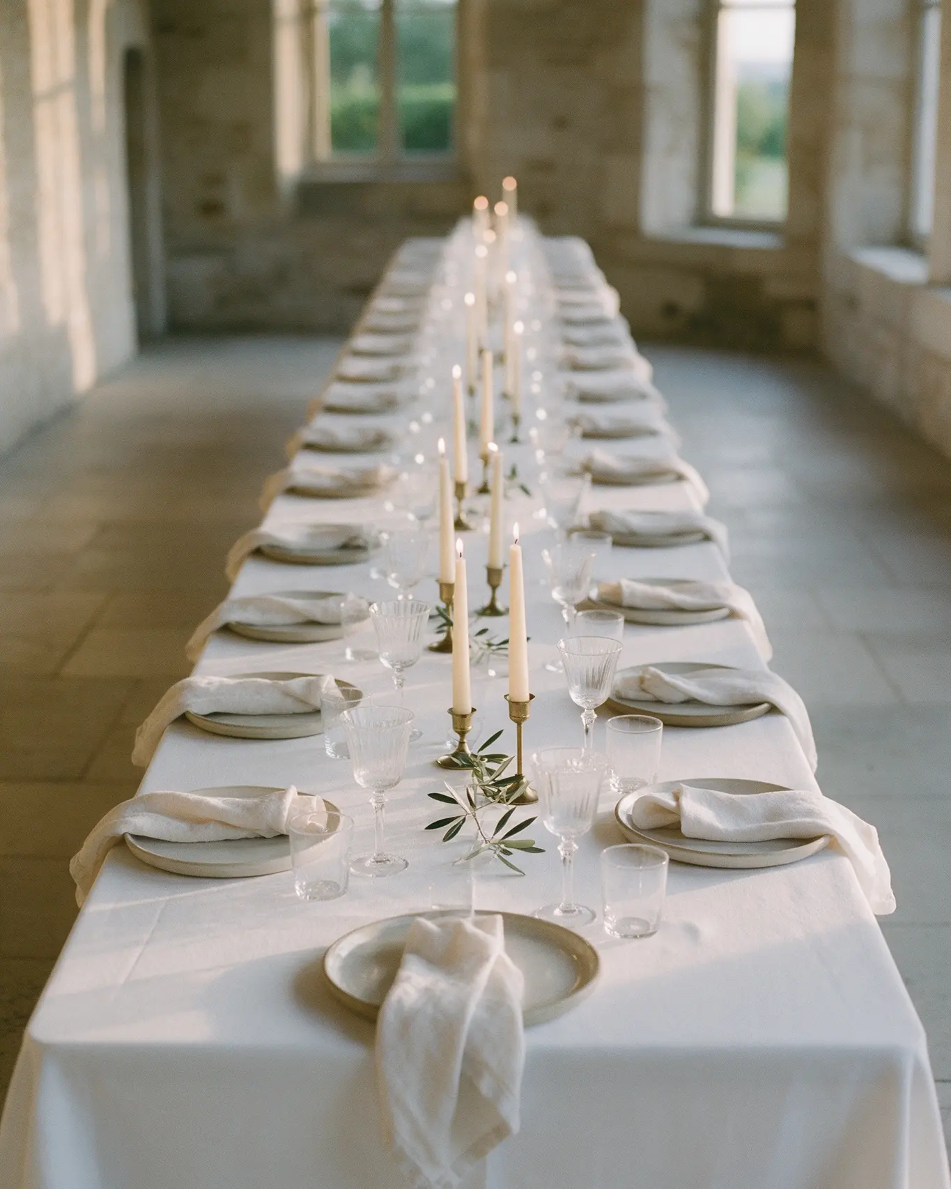 Longue table de réception nappée de blanc, vaisselle en grès, verrerie et bougies sur chandeliers en laiton