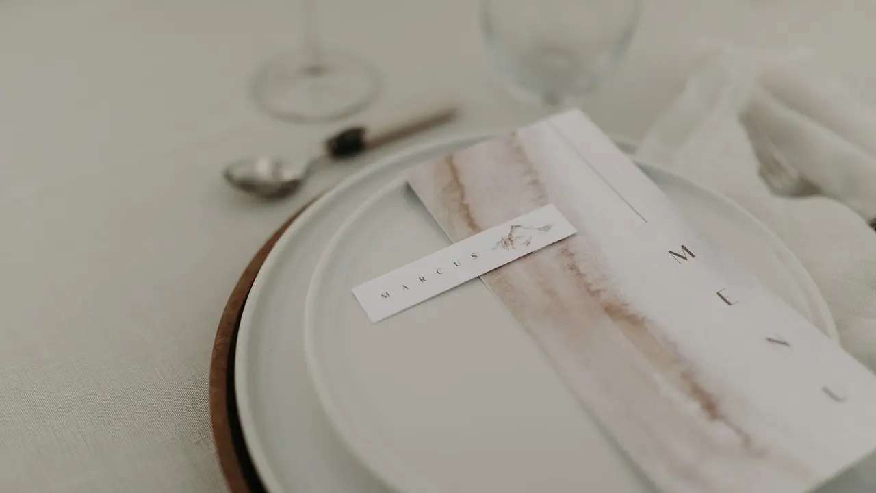 Menu aquarellé posé sur une assiette blanche à liseré bois, nappe en lin
