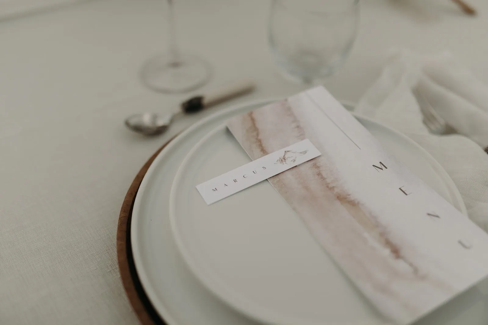 Menu aquarellé posé sur une assiette blanche à liseré bois, nappe en lin